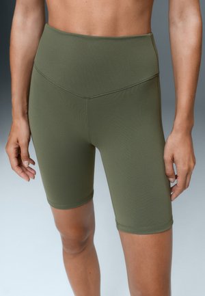 Femme portant un short cycliste taille haute vert olive, debout, mains détendues le long du corps, sur un fond uni.