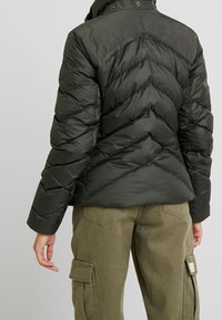 Veste matelassée vert foncé avec un design matelassé, des coutures visibles, un col montant et des boutons-pression. Associée à un pantalon cargo vert olive.