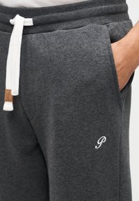 Γκρι φόρμα από απαλό ύφασμα, με κορδόνι στο λευκό, πλαϊνές τσέπες και μικρό λευκό κεντημένο λογότυπο.