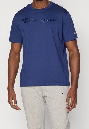 Mężczyzna w granatowym t-shircie Champion z haftowanym logo oraz jasnoszarych spodniach dresowych Champion z małym logo na udzie.