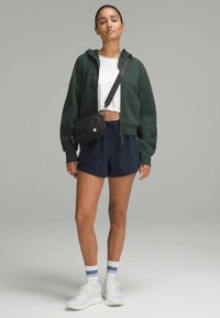 Zelená zkrácená mikina s kapucí, bílý tričko, námořnické šortky, černá crossbody taška, bílé tenisky s modro pruhovanými ponožkami. Neformální sportovní outfit.