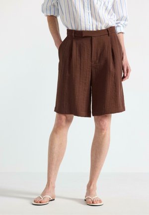 Person iført brune knælange plisserede shorts, en hvid og blåstribet skjorte og hvide sandaler, stående med hænderne i lommerne.