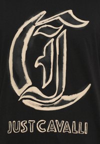 Μαύρη μπλούζα T-shirt με μεγάλο, στυλιζαρισμένο λευκό λογότυπο με σπείρες και καμπύλες, και το κείμενο "JUST CAVALLI" σε λευκό από κάτω.