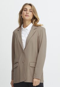 Beige blézer, kerekített hajtókkal, egy gombos záródással és két elülső zsebbel. Fehér inggel párosítva sima textúrával rendelkezik.