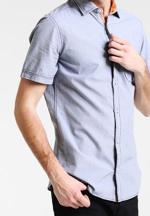 Homme portant une chemise à manches courtes bleu clair à boutons avec un détail orange sur le col, en train d'ajuster le bouton supérieur, sur un fond blanc uni.