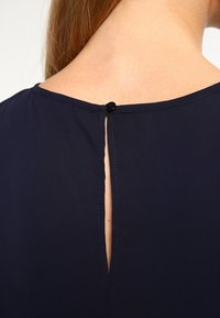 Blouse marine avec un col rond et une ouverture en forme de goutte d'eau au dos, fermée par un bouton noir, en tissu lisse.