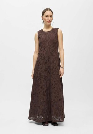 Femme portant une robe longue marron foncé sans manches avec broderie ajourée, debout devant un fond blanc uni.