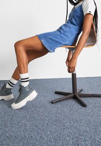 Robe en jean bleue, courte avec des boutons, associée à des baskets montantes grises et des chaussettes rayées. Modèle assis sur une chaise en bois.