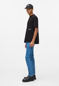 Melna īsām piedurknēm T-kreklu ar brīvu piegriezumu, ar ziliem akcentiem, zilām džinsām un melnām šņorējām kurpēm, stāvot uz balta fona.