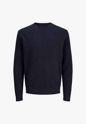 Blond chłopiec stoi, ubrany w granatowy sweter z dzianiny na jasnoniebieskiej koszuli z kołnierzykiem, czarne spodnie i czarne buty, patrząc w bok.