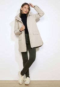 Vrouw draagt een beige knie-lang jack, zwarte coltrui, donkergroene broek en witte sneakers, staand tegen een effen witte muur.