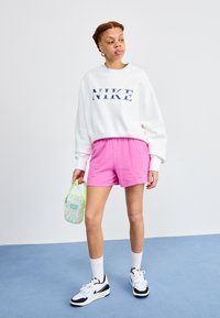 Bijela majica s kapuljačom s plavim Nike logotipom, ružičaste šortse i bijele čarape. Crno-bijele tenisice. Drži malu torbicu s pastelnim uzorkom.