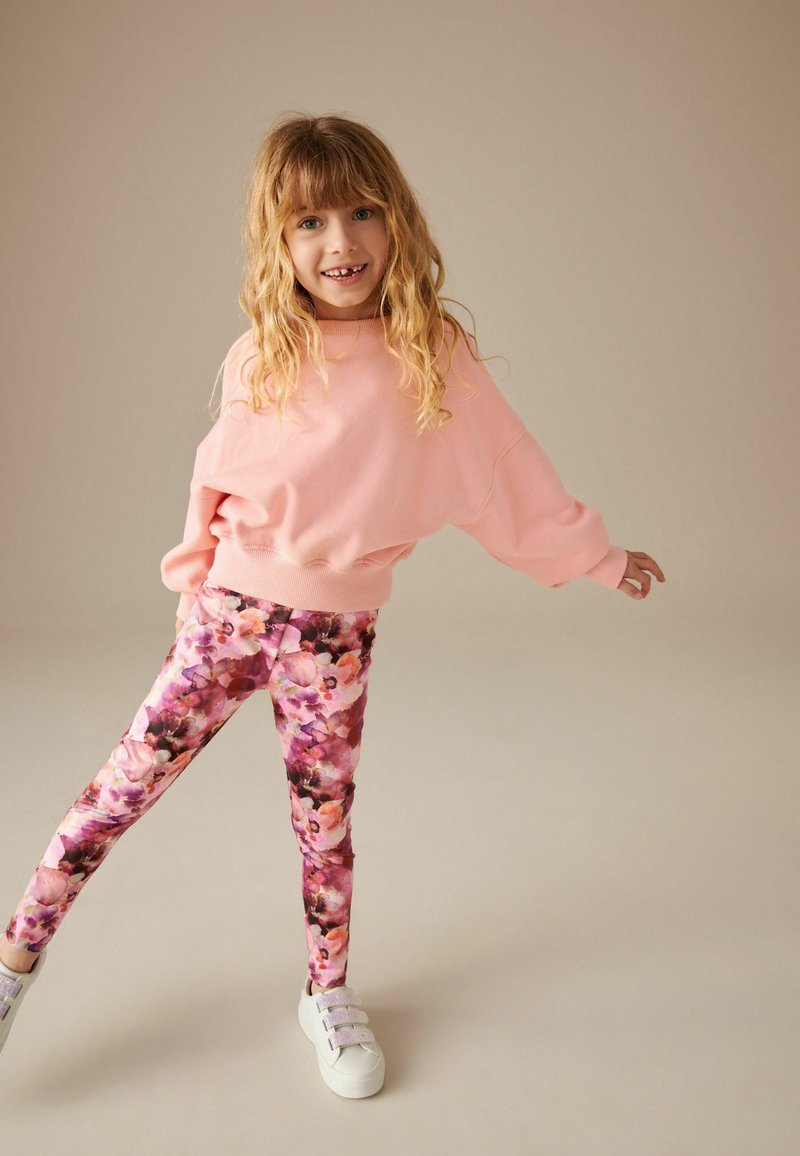 Felpa oversize rosa con maniche lunghe, abbinata a leggings floreali colorati nei toni del rosa, viola e nero. Sneakers bianche.