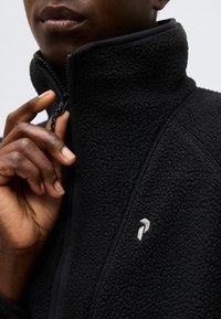 Κοντινό πλάνο ενός ατόμου που κλείνει το φερμουάρ σε μαύρο ζακέτα από φλις με μικρό λευκό κεντημένο λογότυπο στο στήθος.