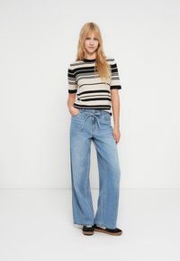 Jeune femme aux longs cheveux blonds portant un t-shirt à manches courtes rayé noir et blanc, un jean large bleu clair et des baskets noires.