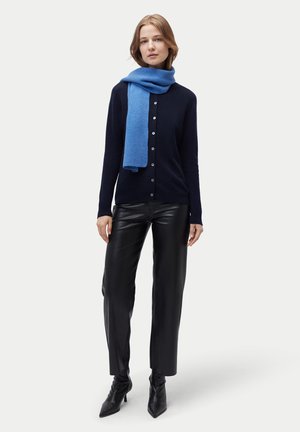 Frau trägt einen schwarzen, geknöpften Cardigan, blauen Schal, schwarze Lederhose und schwarze Stiefel mit hohen Absätzen, steht vor einem schlichten Hintergrund.