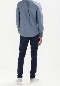 Mężczyzna stoi, ubrany w niebieską koszulę z długim rękawem, ciemne dopasowane spodnie oraz jasnoszare sneakersy z białą podeszwą.