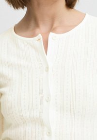 Biały sweter z dzianiny z pionowymi paskami, z okrągłym dekoltem i pięcioma guzikami. Teksturowany wzór z małymi wycięciami w całej powierzchni.