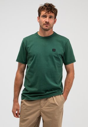 Man met kort krullend haar, draagend een groen T-shirt met korte mouwen en beige broek, staand met één hand in de zak tegen een witte achtergrond.