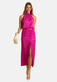 Ensemble deux pièces à motifs floraux roses, comprenant un top à encolure halter et une jupe à fente haute, complété par un sac en osier et des talons beiges à lanières.