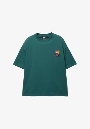 Vihreä puuvillainen ylisuuri t-paita, jossa on pyöreä kaula-aukko ja lyhyet hihat. Vasemmassa rinnassa on pieni brodeerattu peliläppäri-kuvio.