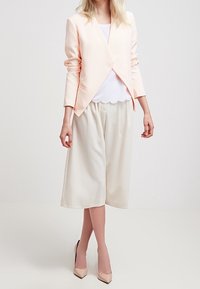 Jasnoróżowa, skrojona marynarka na białej bluzce w kształcie fal, zestawiona z kremowymi, szerokimi spodniami culottes i cielistymi, spiczastymi szpilkami. Gładkie tekstury w całej stylizacji.