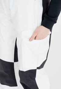 Witte en zwarte bretels met een rits aan de voorkant, grote zijzak en contrasterende kleurblokken in een gladde, matte stof.
