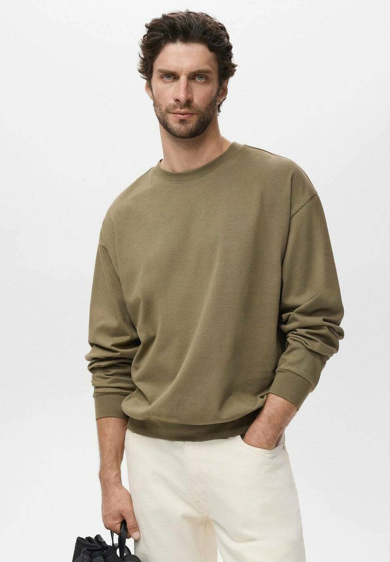Olijfgroene sweatshirt met een ronde hals, geribbelde manchetten en zoom. Model draagt lichtcrème broek en houdt een zwarte tas vast.