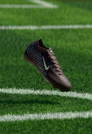 Crampon de football Nike brun foncé avec des points blancs et un logo flottant au-dessus d'un terrain en herbe verte près des lignes blanches de délimitation.