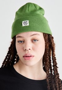 Green knit beanie with a ribbed texture and a small woven label featuring a black logo. The beanie is styled with a slight slouch at the top.