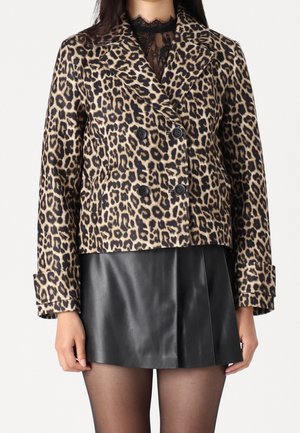 Femme portant une veste à double boutonnage imprimé léopard, un top en dentelle noire, une mini-jupe en cuir noire et des collants noirs transparents sur un fond uni.