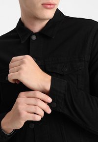 Svart denimjacka med klassisk krage, framfickor och tryckknapps-stängning. Har en strukturerad, hållbar tyg.