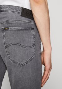 Grå denimjeans med en distinkt fickdesign. Har en svart midjebandsetikett och sömnadsdetalj på fickorna.