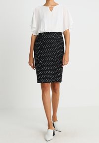 Blusa bianca con maniche a farfalla e dettaglio a chiave, abbinata a una gonna nera aderente a pois. La modella indossa scarpe bianche con apertura sul retro.