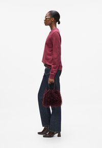 Bordeauxrote Sweatshirt, blaue weite Jeans, braune Lack-Ankelstiefel und eine strukturierte bordeauxfarbene Felltasche. Das Model steht im Profil vor einem weißen Hintergrund.