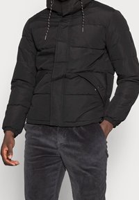 Giubbotto puffa nero con cappuccio, caratterizzato da un design trapuntato, polsini elastici e una tasca sul petto. Il tessuto appare liscio e leggero.