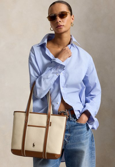 Femme portant des lunettes de soleil, chemise oversize bleu clair, jean bleu, boucles d'oreilles créoles dorées, tenant un sac cabas en cuir beige et marron avec logo.