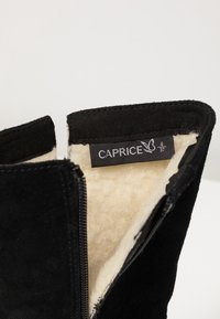 Black suede ankle boot with a cream-colored fur lining, side zipper, and a label reading "CAPRICE" inside the opening. Size 37.