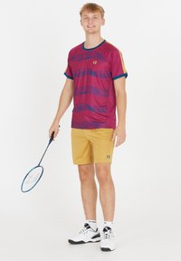 Bordåbne sports T-shirt med blå accenter og stribet mønster, parret med gyldne shorts. Holder en badmintonketsjer og har sorte og hvide sko på.