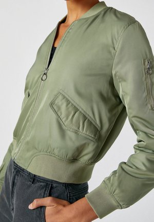 Person trägt eine hellgrüne Bomberjacke mit Reißverschluss, schräger Klappentasche und Reißverschlusstasche am Ärmel, kombiniert mit einer schwarzen Hose.