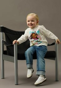 Uśmiechnięte małe dziecko z blond włosami, ubrane w bluzę z postacią z kreskówki, dżinsy i białe trampki, siedzi na czarnym krześle na szarym tle.