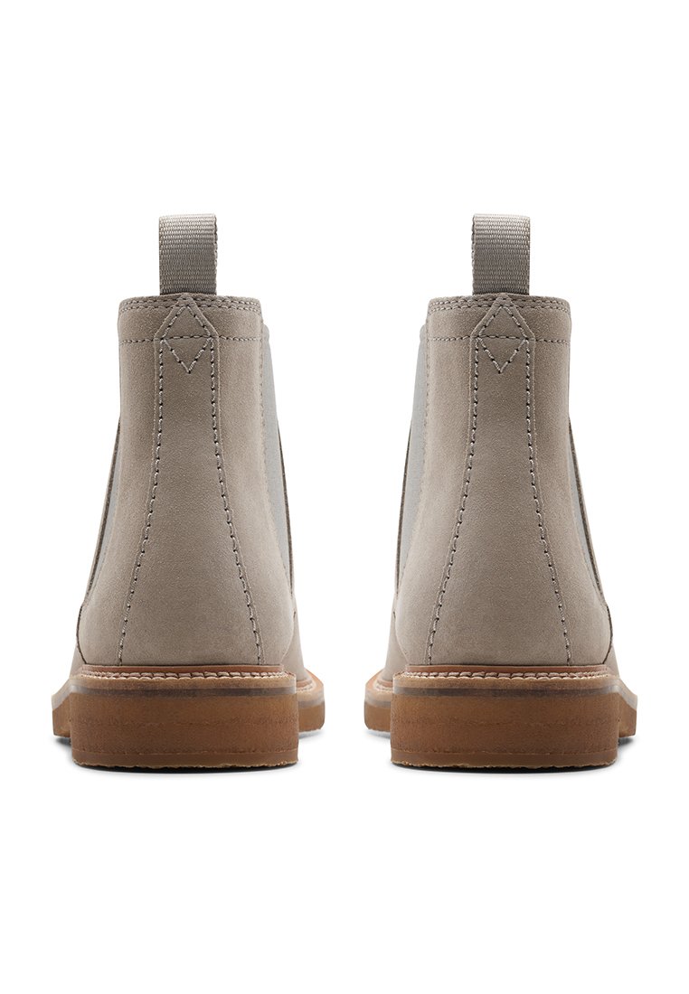 Ein Paar hellbeige Wildleder-Stiefeletten mit braunen Sohlen, sichtbaren Nähten und Zuglaschen hinten, von hinten auf einem weißen Hintergrund gezeigt.