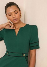 Femme portant une robe verte avec des bordures noires et des accents en bouton doré, posant avec une main près du cou et des boucles d'oreilles dorées en statement.