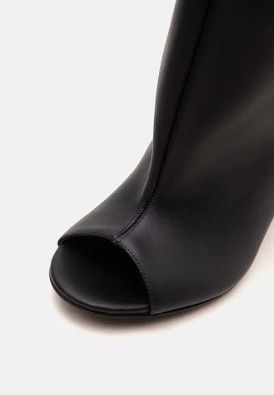 Bottine en cuir noir à bout ouvert avec couture avant visible et texture lisse sur fond blanc.