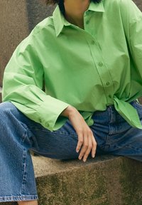 Camisa verde de botón con mangas largas y abullonadas, atada en la cintura. Combinada con jeans de mezclilla azul claro, sentada sobre una superficie de piedra.