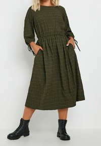Tummanvihreä ruutukuvioinen mekko, jossa on pitkät hihat ja korostettu vyötärö; sisältää taskut ja nauhakoristeet. Yhdistetty mustiin maiharikenkiin.