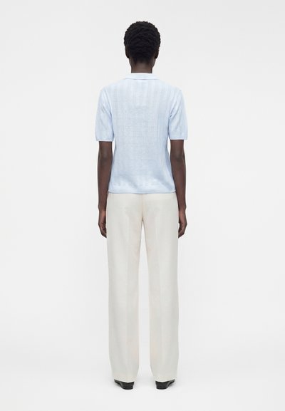 Personne aux cheveux courts et bouclés portant un haut en maille bleu clair à manches courtes et un pantalon blanc ample, debout de dos sur un fond blanc.
