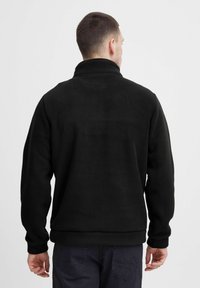 Svart fleecejakke med høy krage, lange ermer og ribbestrikkede mansjetter. Jevn tekstur med minimal detaljering, laget for komfort.