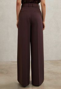 Plaša silueta, augstas jostas bikses tumši vīnsarkanā audumā. Raksturo gluda tekstūra un divas aizmugurējās kabatas, izstrādātas ar piegriezumu pēc pasūtījuma.