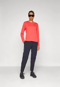 Moteris vilki raudoną ilgomis rankovėmis marškinį, juodas treniruotes kelnes, juodus sportinius batelius ir dideles juodas saulės akinius, stovinti prieš pilką vientisą foną.